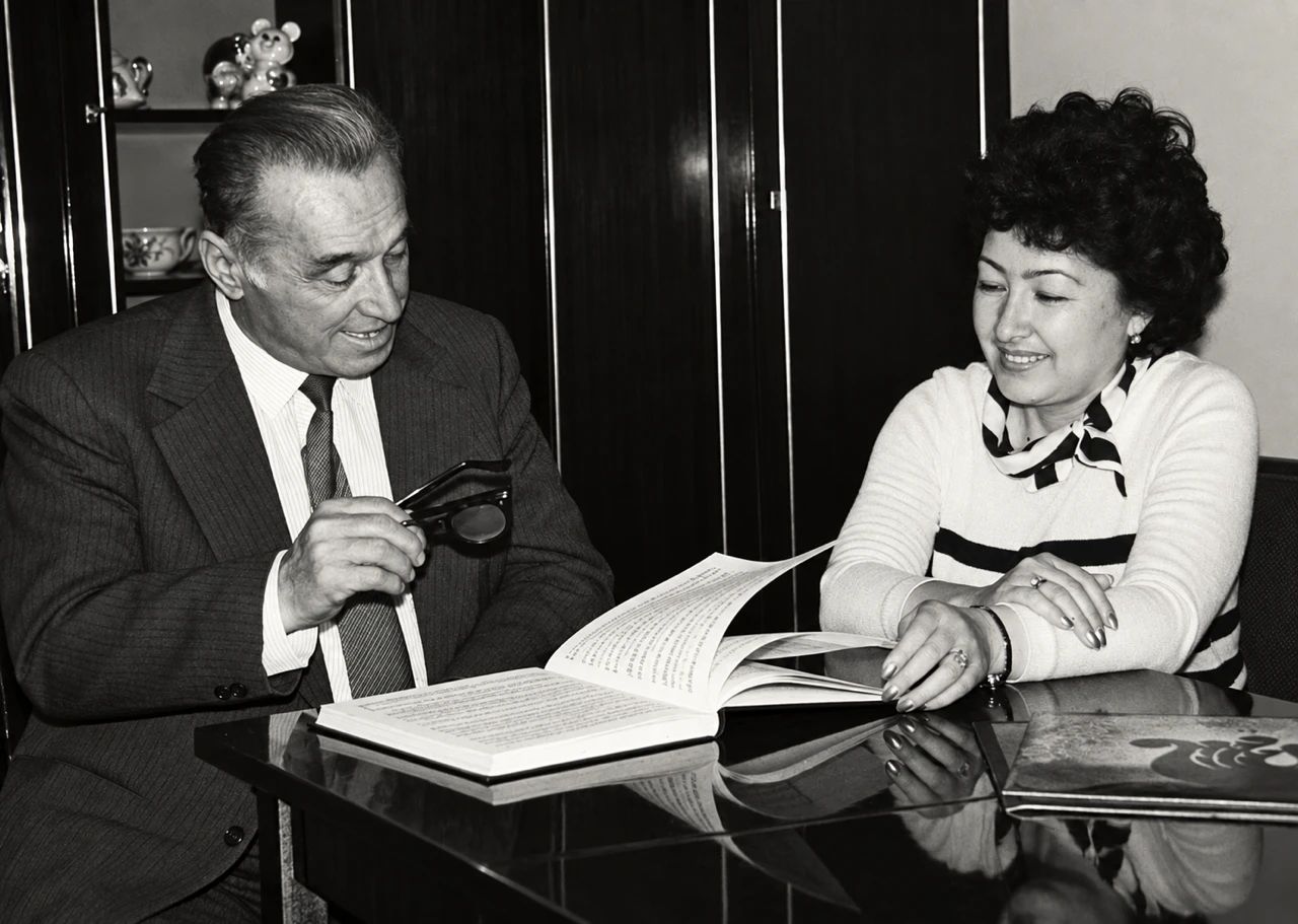 Black-and-white photograph of Ziyodullo Shahidi, holding his glasses, and Munira Shahidi, smiling, at a polished table with a large open book between them.